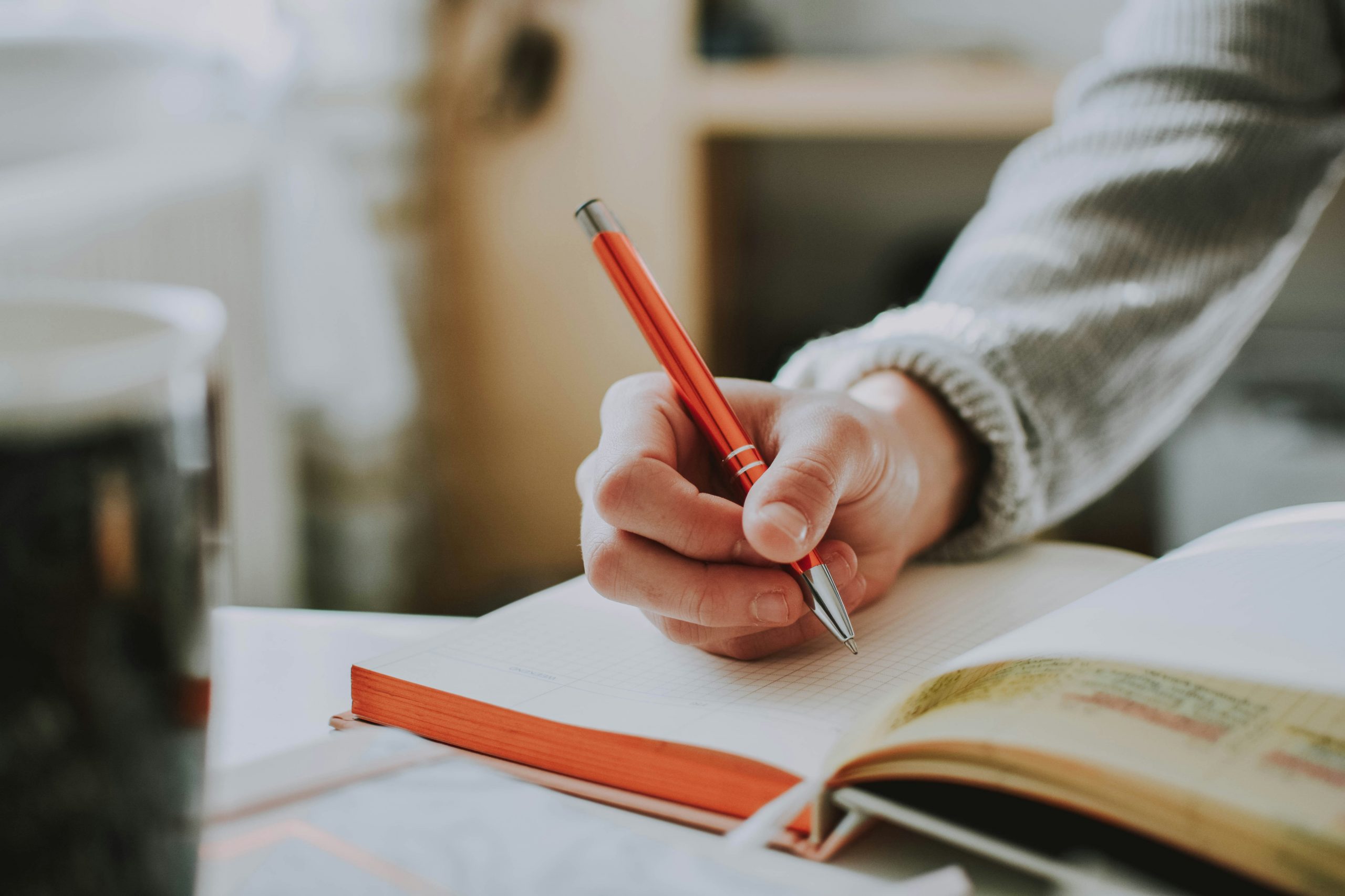 Student writing a scholarship recommendation request in a notebook with a red pen; open book and mug on the desk.